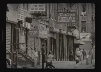 A grainy black and white photo of Italian East Harlem in the 1950s-1960s featured a sidewalk with various eateries and shops and kids playing.
