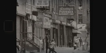 A grainy black and white photo of Italian East Harlem in the 1950s-1960s featured a sidewalk with various eateries and shops and kids playing.